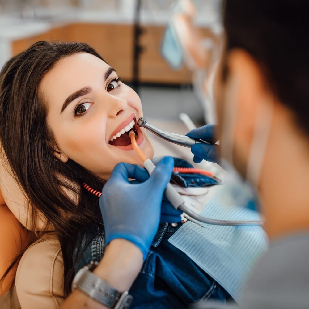 young-female-patient-visiting-dentist-office Young female patient visiting dentist office. Beautiful woman with healthy straight white teeth sitting at dental chair with open mouth during oral checkup while doctor working at teeth.